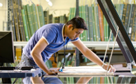 a man leaning over a scanner on a waist height desk holding a document in place before closing the scanner cover, a substantial number of large books are visible on shelves in the background