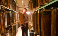 A man standing between two sets of shelves each containing numerous volumes of newspapers bound into large books, the man is holding and examining one of the volumes.